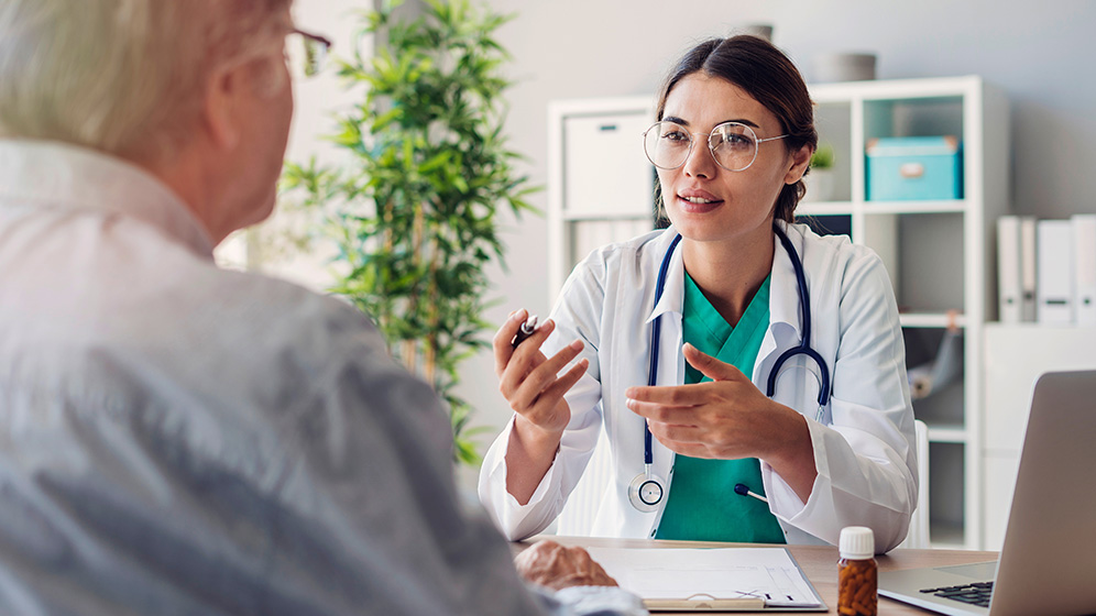 Junge Ärztin mit Brille spricht in einem Behandlungszimmer mit einem Patienten.