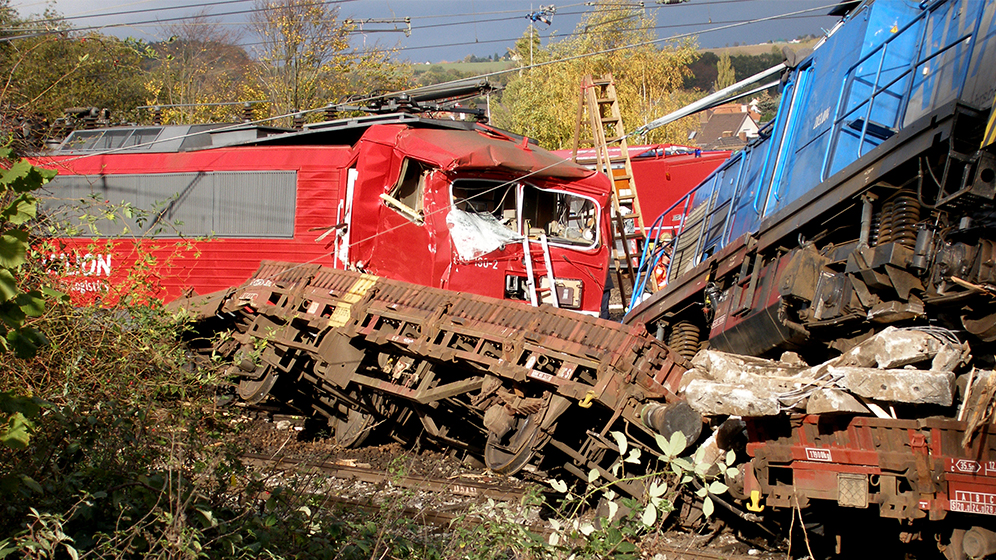 Zugunfall mit stark beschädigten Lokomotiven und Waggons auf den Gleisen, umgeben von Vegetation und Rettungsarbeiten im Hintergrund.