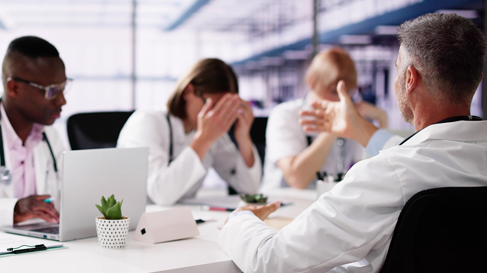 Ärzte in einem Besprechungsraum, einer hält den Kopf in den Händen, während ein anderer spricht; Laptop und Notizen auf dem Tisch.
