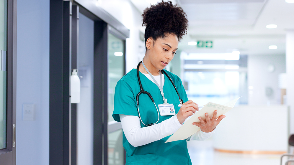 Medizinisches Fachpersonal in grüner Arbeitskleidung steht in einem Krankenhausflur und überprüft Dokumente, mit einem Stethoskop um den Hals.