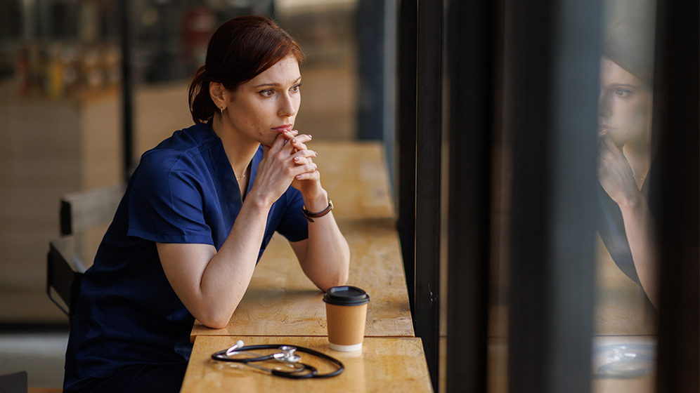 Frau in blauer, medizinischer Arbeitskleidung sitzt nachdenklich an einem Holztisch mit einem Kaffeebecher und einem Stethoskop.