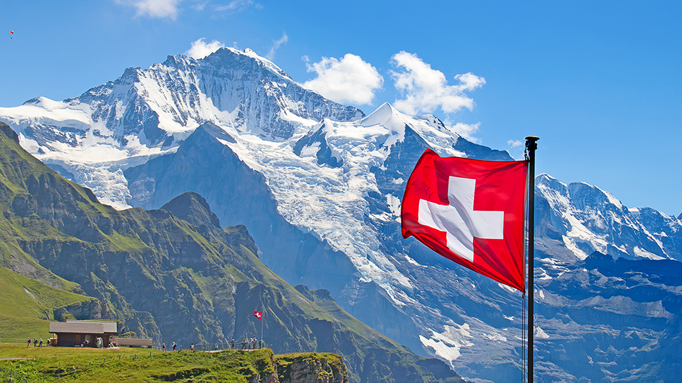 Schweizer Flagge vor einer beeindruckenden Berglandschaft mit schneebedeckten Gipfeln und grünem Tal unter klarem, blauem Himmel.